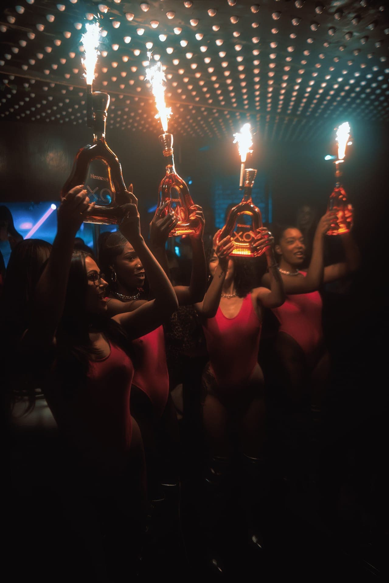 Guests in a dark nightclub holding up bottles topped with lit sparklers under a ceiling of small lights