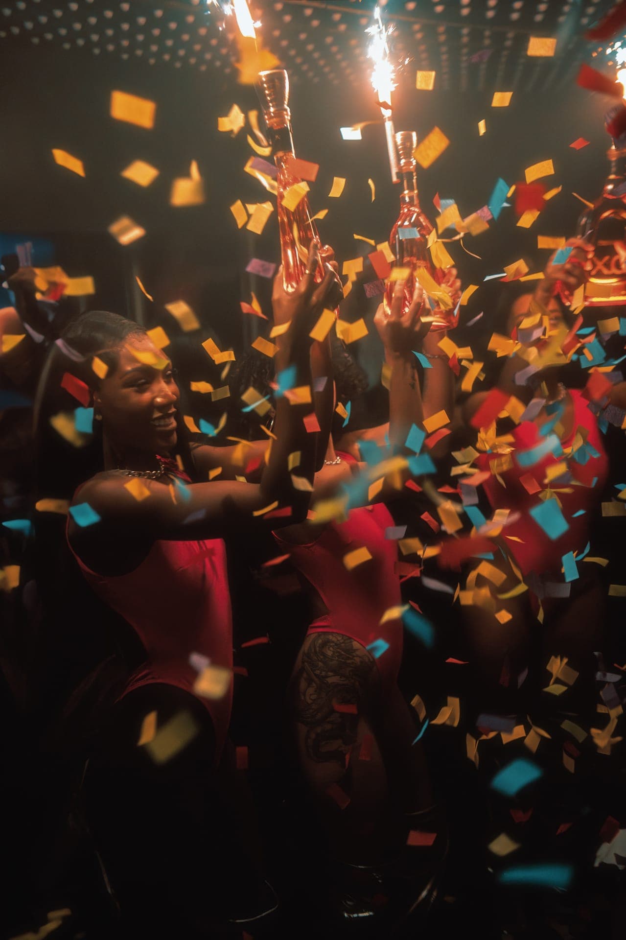 Smiling guests holding up bottles topped with sparklers as colourful confetti falls in a dimly lit club