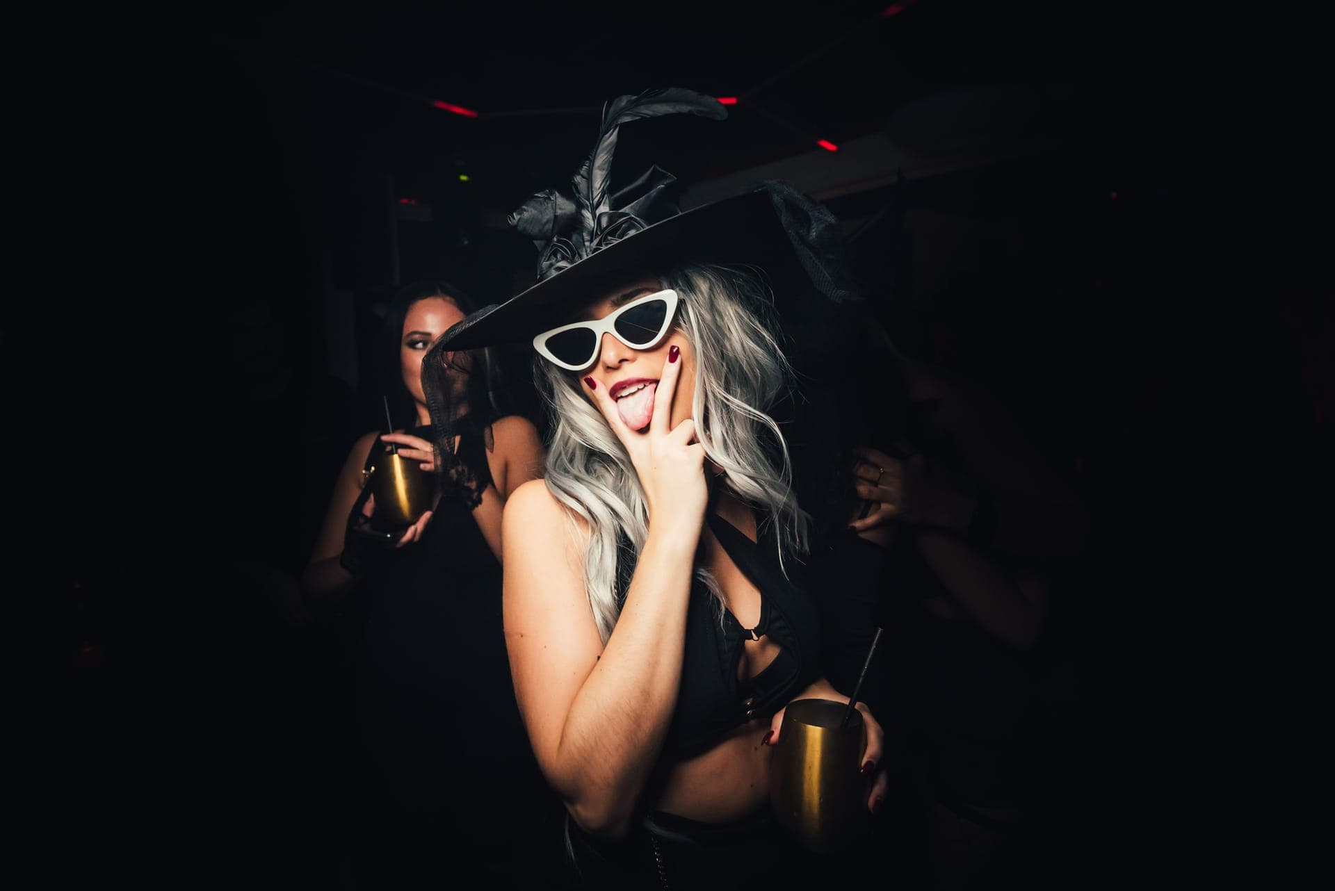 A woman in a feathered black witch hat and white cat-eye sunglasses holding a gold cup on a dark dance floor